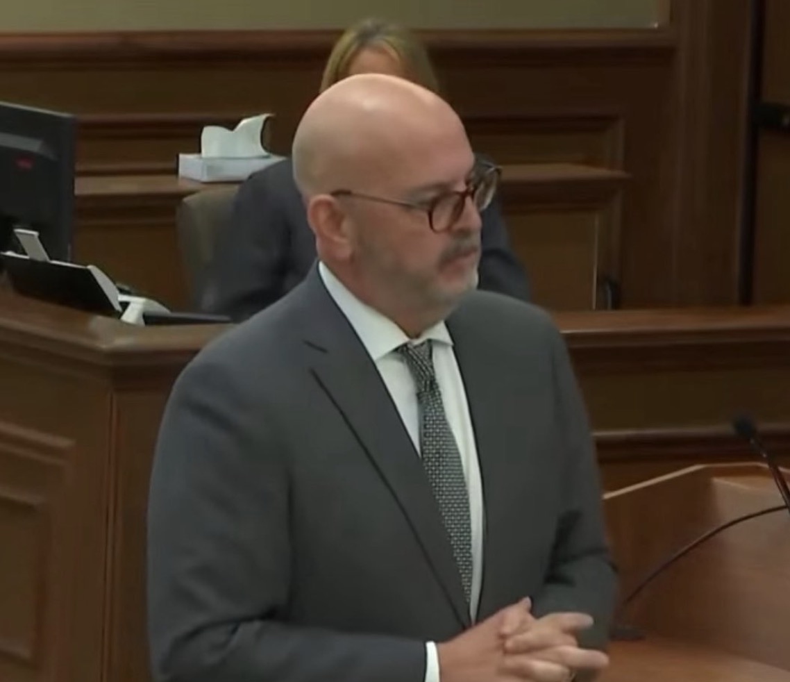Douglas Weinstein speaking at a courtroom podium in a gray suit